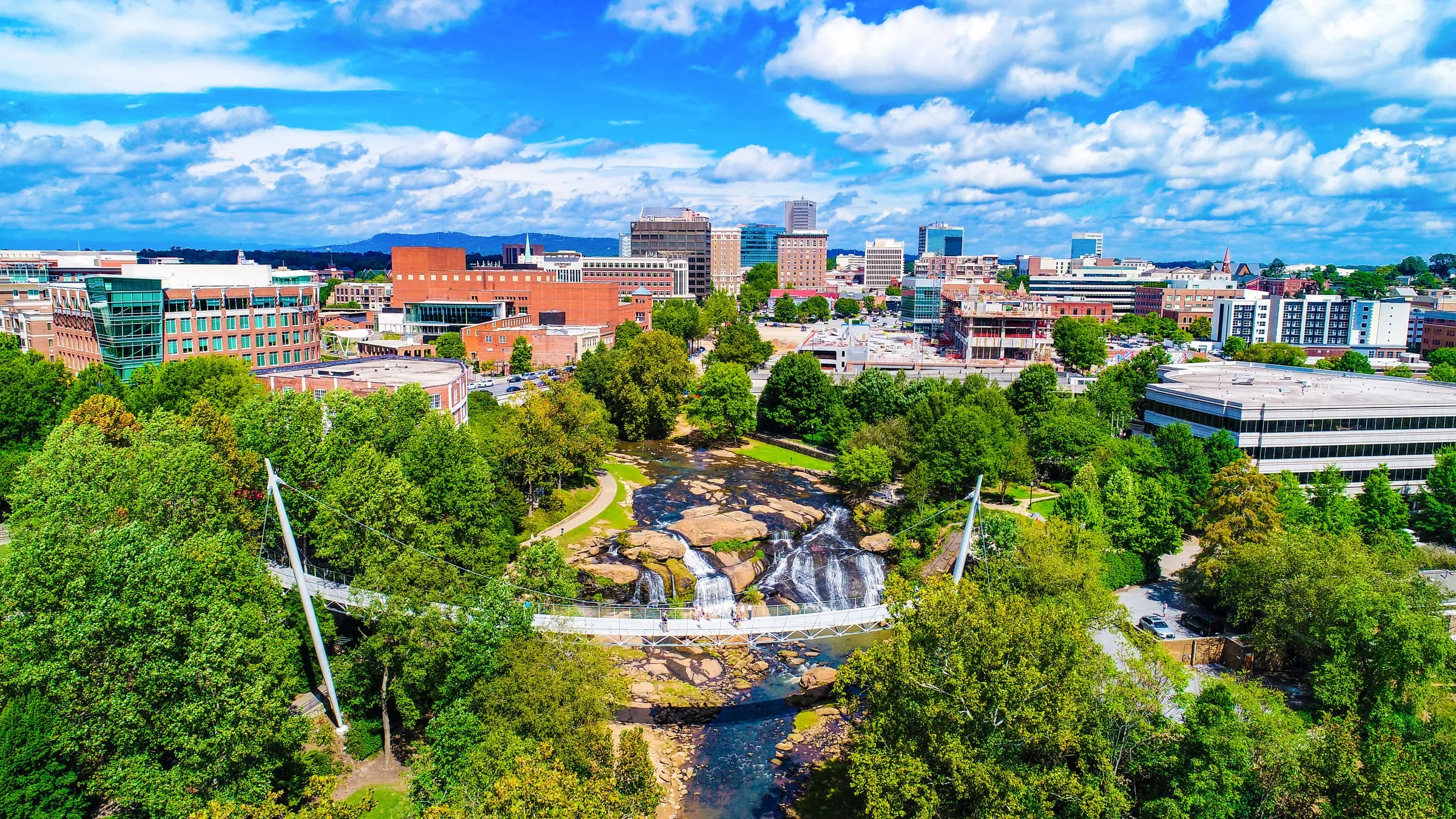 Aerial view of Falls Park on the Reedy in downtown Greenville, South Carolina, with the Liberty Bridge crossing the river above the falls and the city skyline behind.